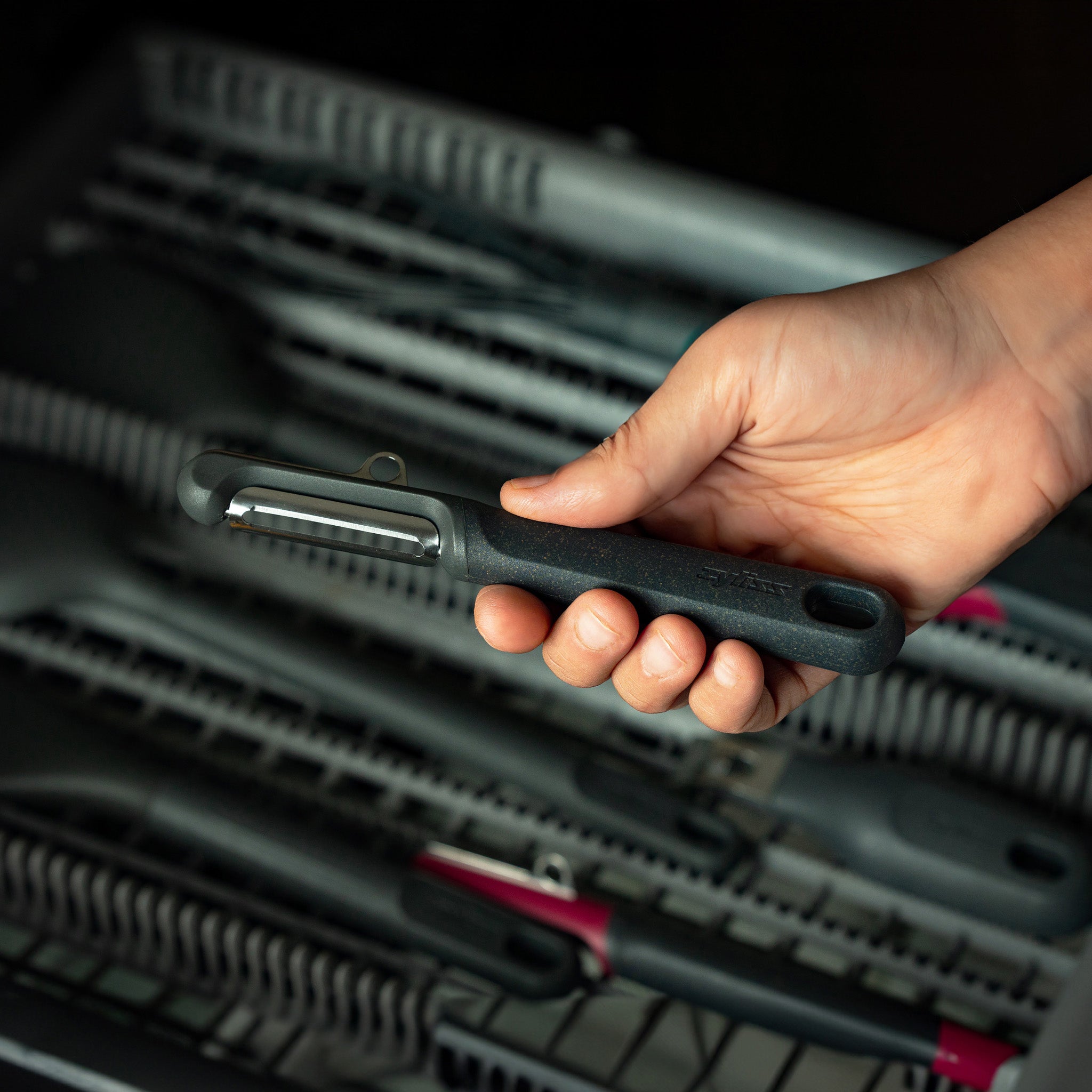 A hand holding a black vegetable peeler above the open rack of a dishwasher filled with other kitchen utensils.
