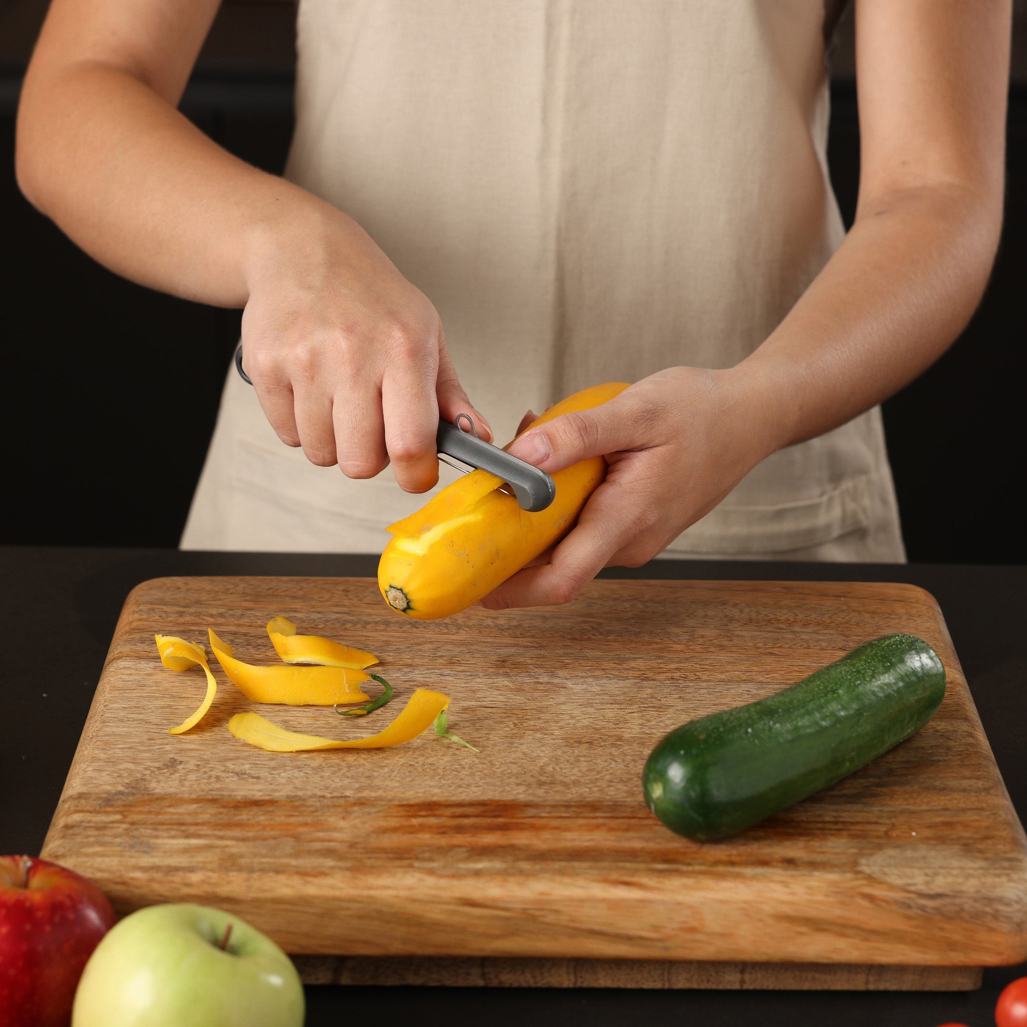 A person wearing a beige apron is peeling a yellow squash with a peeler over a wooden cutting board, which also holds a green zucchini, an apple, and a green fruit.