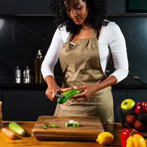 A woman in a beige apron uses the Zyliss Swivel Peeler with an ergonomic handle to peel a cucumber in a modern kitchen. Apples, lemons, and other vegetables are arranged on the wooden counter beside her. 
