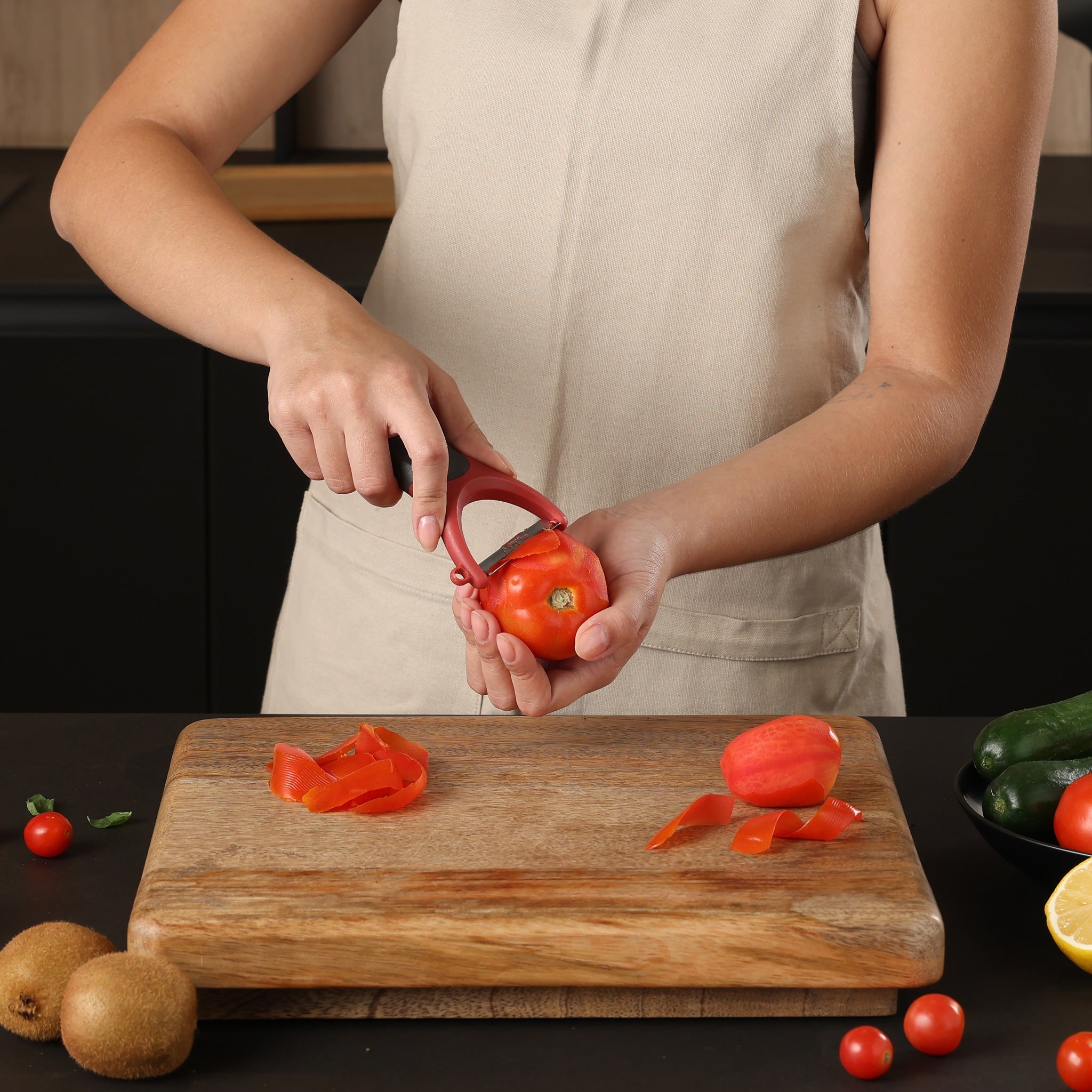 Wearing a beige apron, someone peels a red bell pepper with the Zyliss Soft Skin Y Peeler, featuring an ergonomic grip, over a wooden cutting board on an eco-friendly dark kitchen counter with assorted fruits and vegetables.