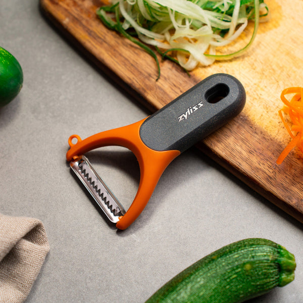 The Zyliss Julienne Y Peeler, featuring an ergonomic orange and black handle, rests on a gray countertop near a wooden board with shredded cucumber and carrot—an essential eco-friendly kitchen tool.