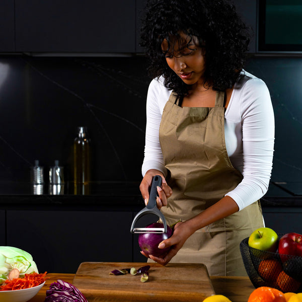 In a modern kitchen, a woman in a beige apron uses the Zyliss Wide Peeler to peel a purple onion on a wooden cutting board, surrounded by bowls of fresh fruits and vegetables.