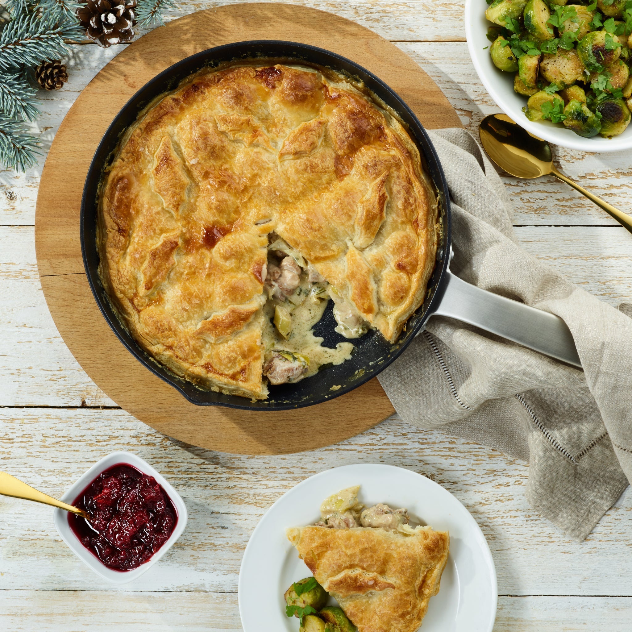 A skillet chicken pot pie with a golden, flaky crust sits on the Zyliss Ultimate Pro Non-Stick Frying Pan With Pouring Lip. A slice is served with Brussels sprouts beside bowls of cranberry sauce and more sprouts on a rustic white table. #28cm/11"