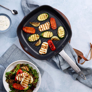 The Zyliss Ultimate Pro Non-Stick Square Grill Pan With Pouring Lip grills halloumi, zucchini, onion, and red pepper slices, shown next to a grilled vegetable salad and creamy dip on a blue background.