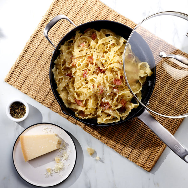 A pan of mafaldine pasta with bacon and pepper sits on a woven mat. Cooked in the Zyliss Ultimate Pro Non-Stick Saute Pan & Glass Lid, made from hard-anodized aluminium and PFOA-free, its ideal for effortless cooking and cleanup. Parmesan and black pepper are nearby.