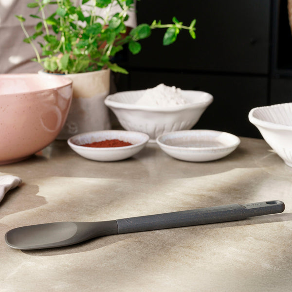 A Zyliss Spoontula with an ergonomic handle lies on a kitchen counter next to bowls of flour, spices, and a green plant, softly lit by sunlight. #Large