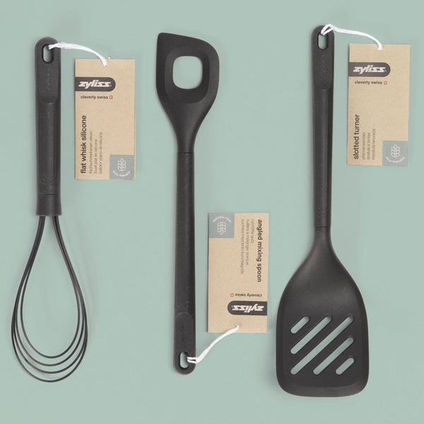 Three black kitchen utensils—a silicone whisk, a mixing spoon with a hole, and a slotted turner—are laid out on a light green background, each with a brown Zyliss brand tag attached.