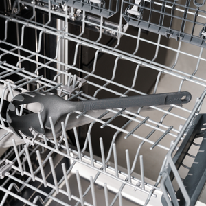 A black spaghetti server utensil is placed inside the bottom rack of an open dishwasher. The dishwasher racks are empty except for the utensil.