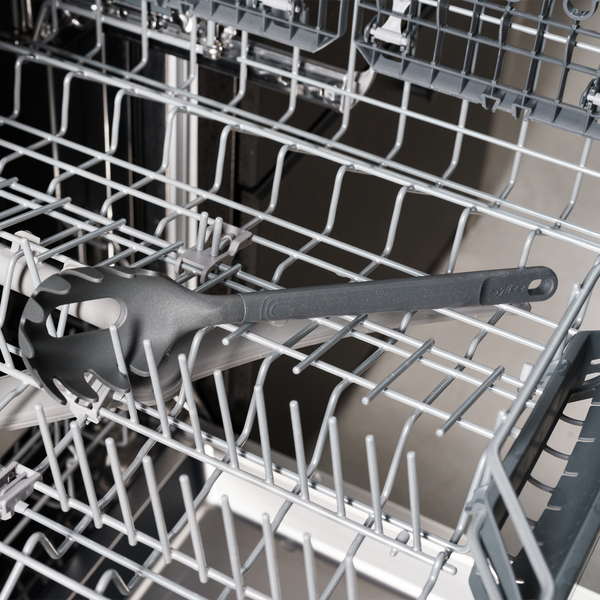 A black spaghetti server utensil is placed inside the bottom rack of an open dishwasher. The dishwasher racks are empty except for the utensil.