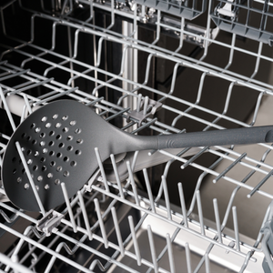 A metal slotted spoon placed in the bottom rack of an open dishwasher, with racks and other compartments visible in the background.