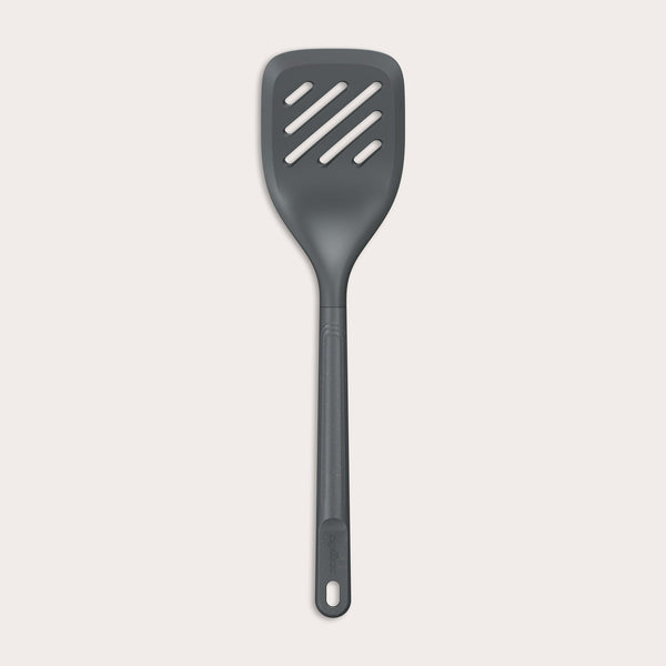 The Zyliss Slotted Turner, shown in black with three diagonal slots and an ergonomic handle, features a heat-resistant design for safe cooking or flipping food. Displayed against a plain white background.