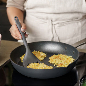 A person wearing a beige apron uses a black spatula to flip potato pancakes cooking in a black frying pan on a stovetop.