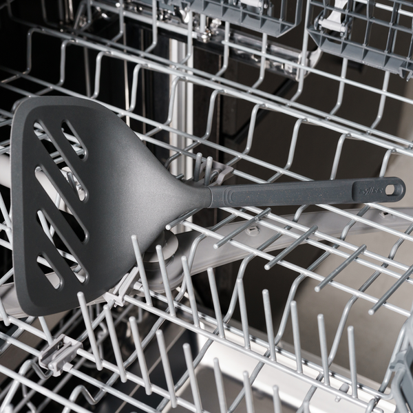 A black slotted spatula rests on the top rack of an open dishwasher, surrounded by metal prongs and other dish compartments.