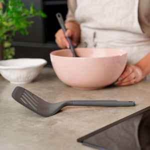 Wearing an apron, a person stirs a pink mixing bowl on the kitchen counter as the Zyliss Turner XL, a heat-resistant turner with an ergonomic handle, rests in the foreground. Also visible are a white bowl and a green plant.