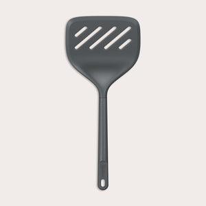 A Zyliss Turner XL slotted spatula in black, featuring a wide square head with three diagonal slots, ergonomic handle, and heat-resistant design is displayed against a plain light background.