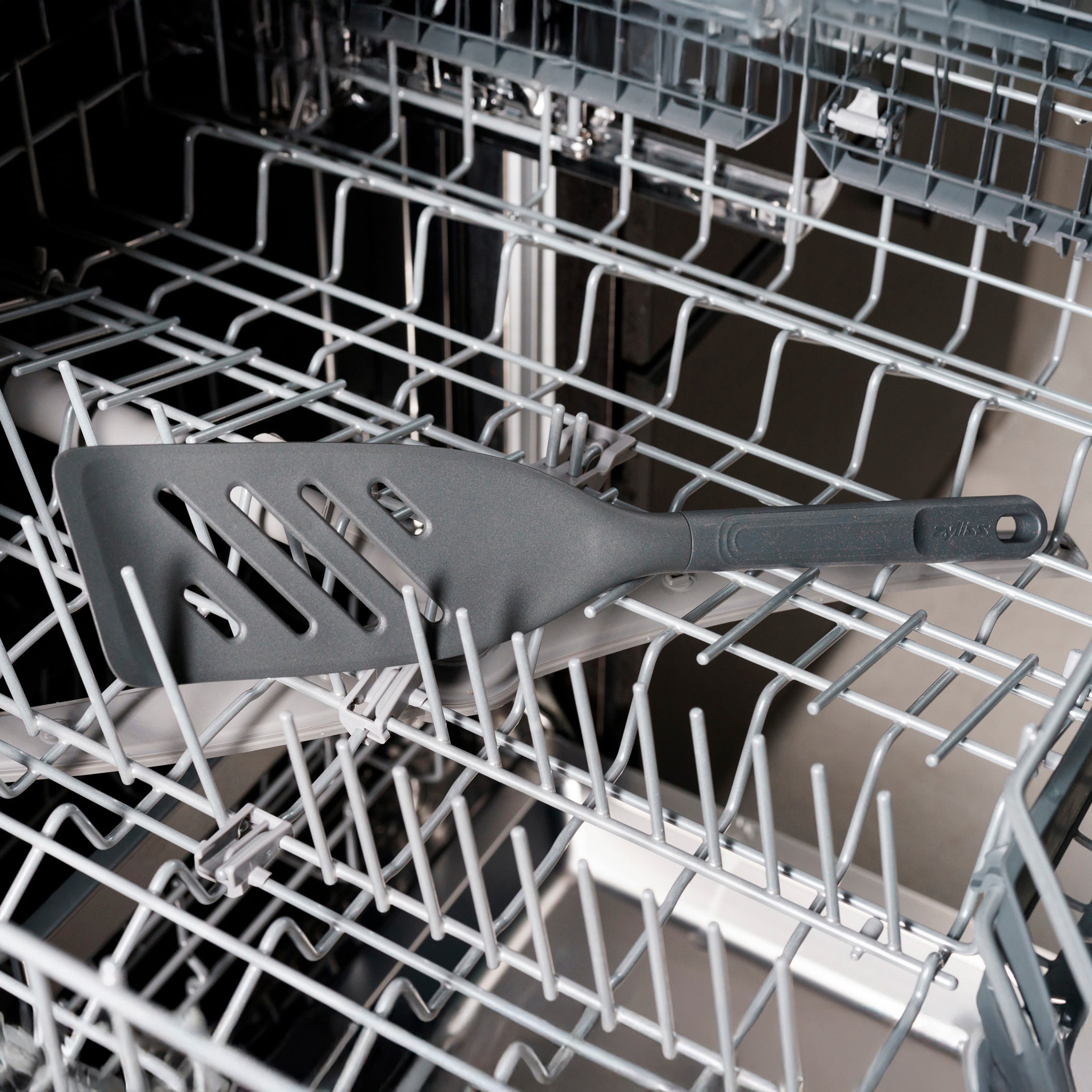 A black slotted spatula placed diagonally on the top rack of an open dishwasher with empty metal prongs visible.