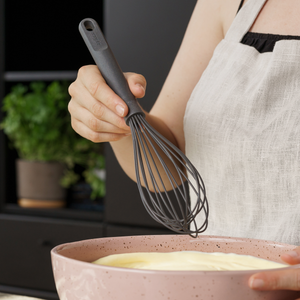 A person wearing a beige apron is whisking batter in a pink speckled bowl. The focus is on their hand holding a black whisk. There is a potted plant blurred in the background.