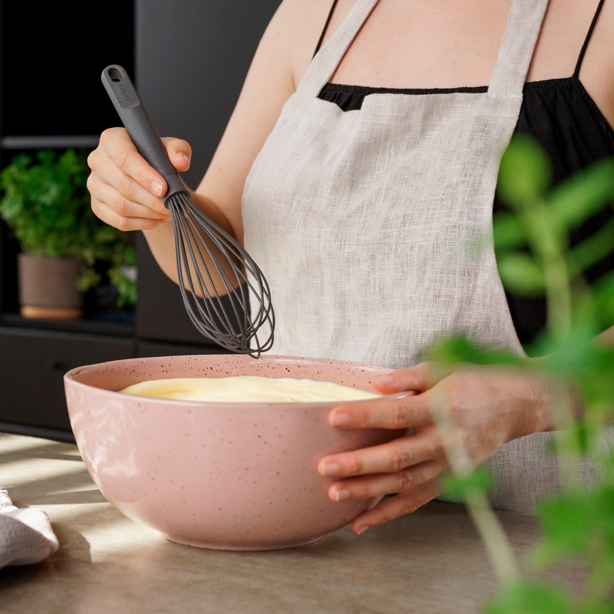 Wearing a linen apron, a person uses the Zyliss Balloon Whisk Silicone to mix batter in a pink bowl in a kitchen with green plants. The non-stick cookware safe whisk is ideal for everyday baking and cooking tasks.