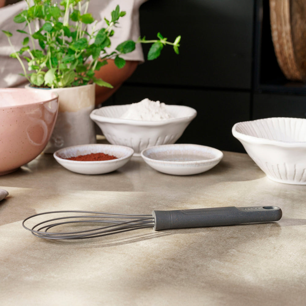 The Zyliss Flat Whisk Silicone features a black, heat-resistant silicone design with a long handle and looped wires, displayed on a plain light background.