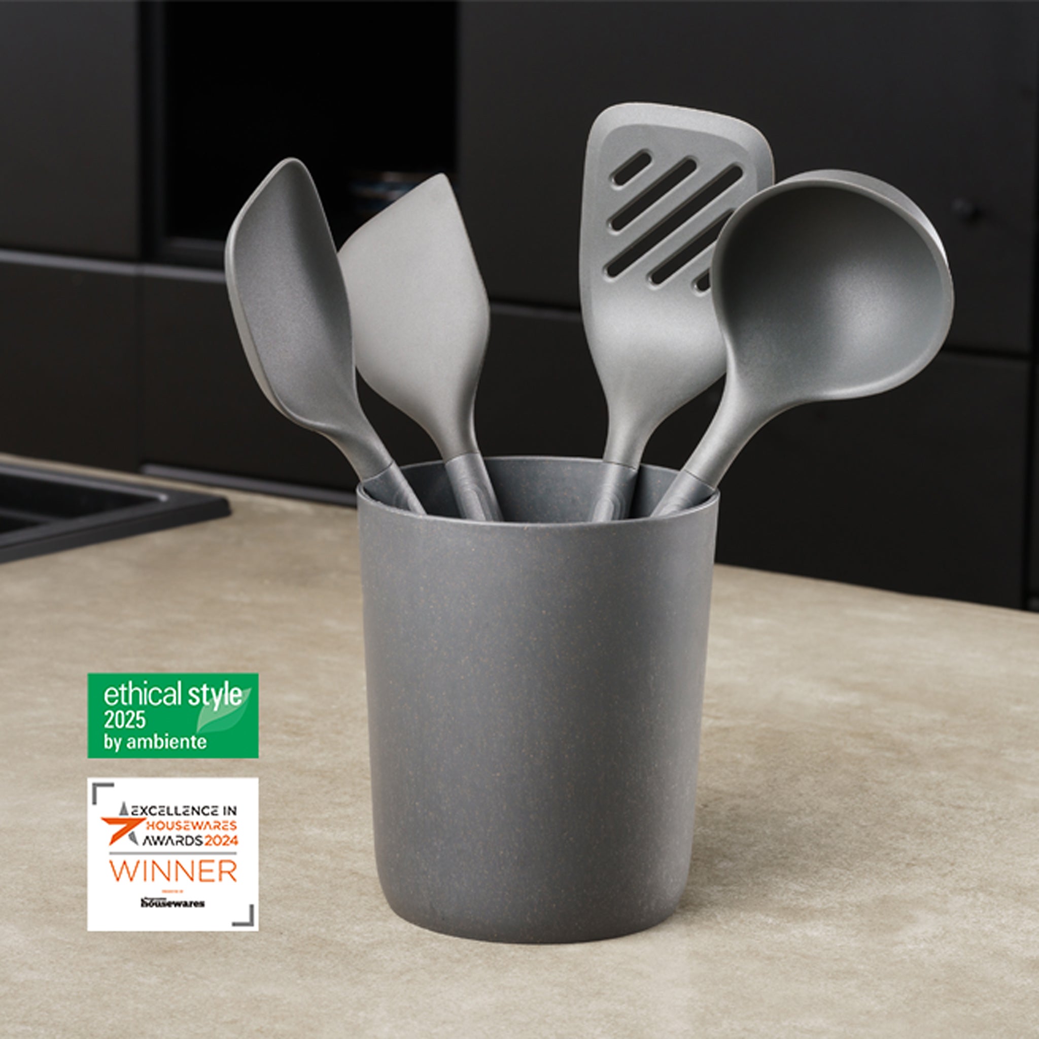 A set of five gray kitchen utensils in a matching holder on a countertop, with two award labels nearby for ethical style (2025) and excellence in business innovation (2024).