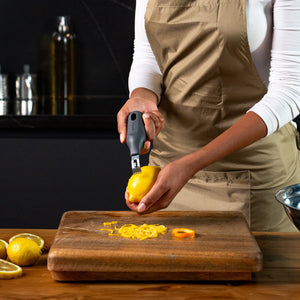 Wearing a beige apron, a person uses the Zyliss 2-In-1 Zester to zest a lemon over a wooden cutting board, with fresh lemon zest and sliced lemons close by on the counter.
