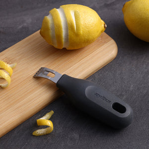 The Zyliss 2-In-1 Zester with a black handle sits on a wooden board beside a partially zested lemon, lemon peel strips, and a whole lemon on a dark surface.