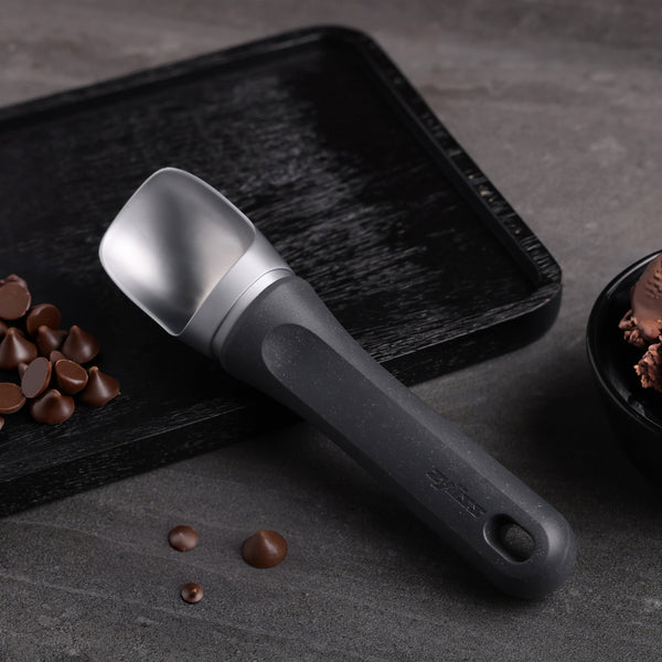 #belugagrey A Zyliss Ice Cream Scoop with an eco-friendly handle sits on a textured black tray beside chocolate chips and a bowl of chocolate ice cream, arranged on a gray surface.