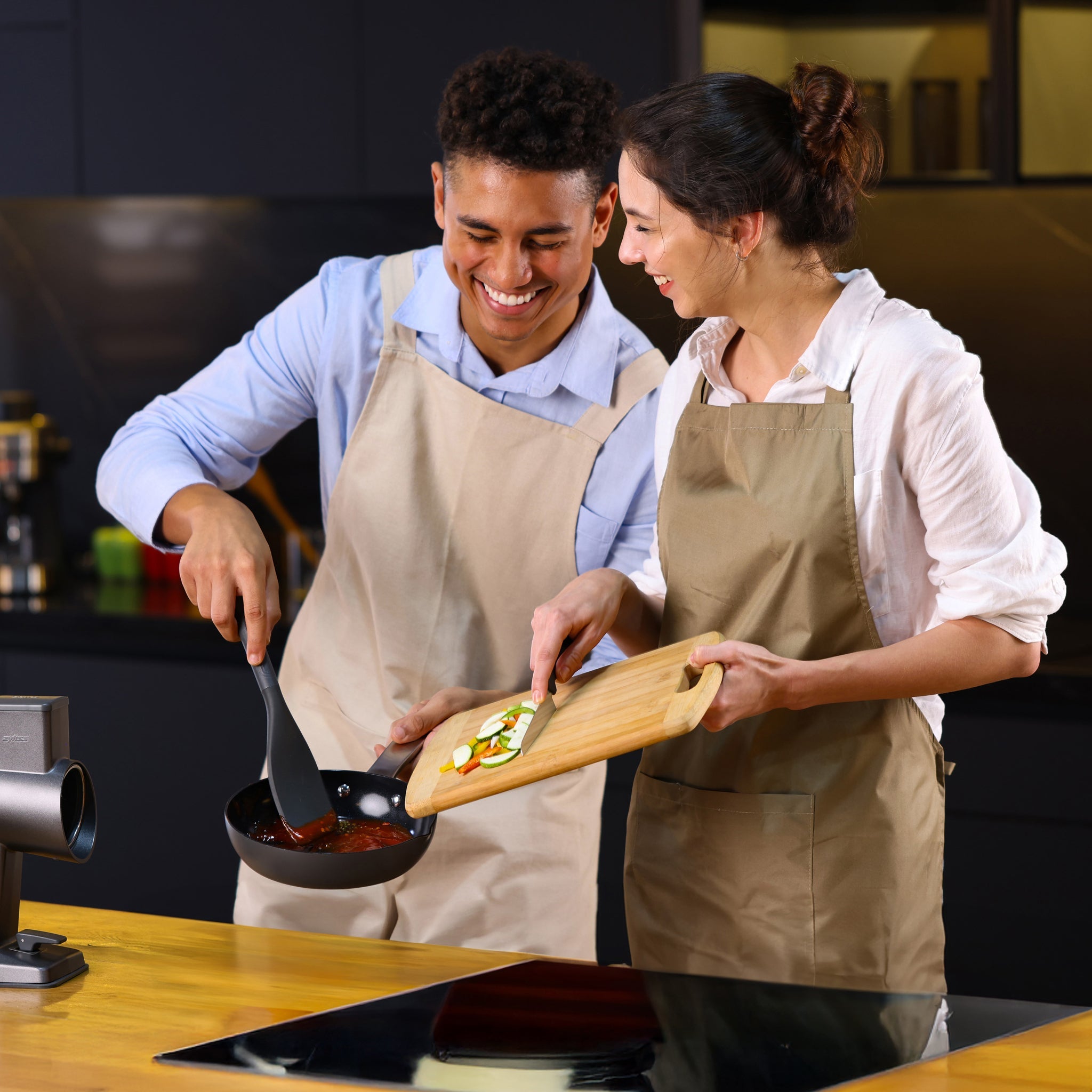 Two people in aprons cook together in a modern kitchen, smiling as one stirs food in the Zyliss Ultimate Pro Ceramic Fry Pan while the other adds chopped vegetables from a wooden cutting board. #20cm/8"