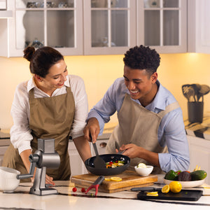 A woman and man in aprons smile and cook together in a modern kitchen. The man stirs food in the Zyliss Ultimate Pro Ceramic Fry Pan while the woman watches. Fresh ingredients and utensils are on the counter. #24cm/9½"