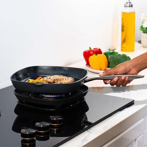 A hand holds a frying pan on a stovetop, cooking seasoned chicken and vegetables. Colorful bell peppers, broccoli, and a bottle of oil are on the white counter in the background.