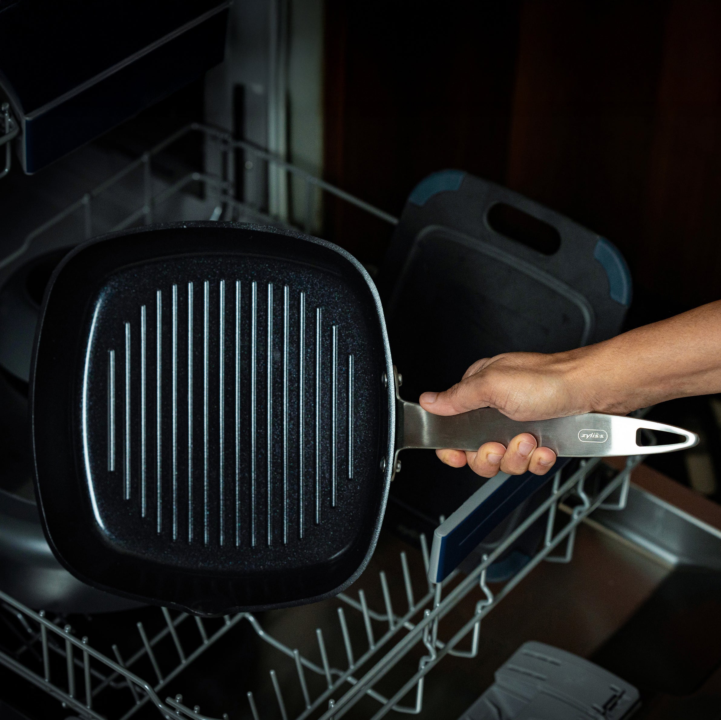 A hand places a black grill pan with a metal handle into the bottom rack of an open dishwasher. Other kitchen items are visible inside the dishwasher.