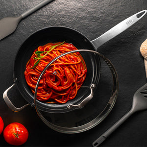 A pot of cooked spaghetti in rich tomato sauce, garnished with a sprig of rosemary, sits on a dark surface with a glass lid partially covering it. Two spatulas and fresh tomatoes are placed nearby.