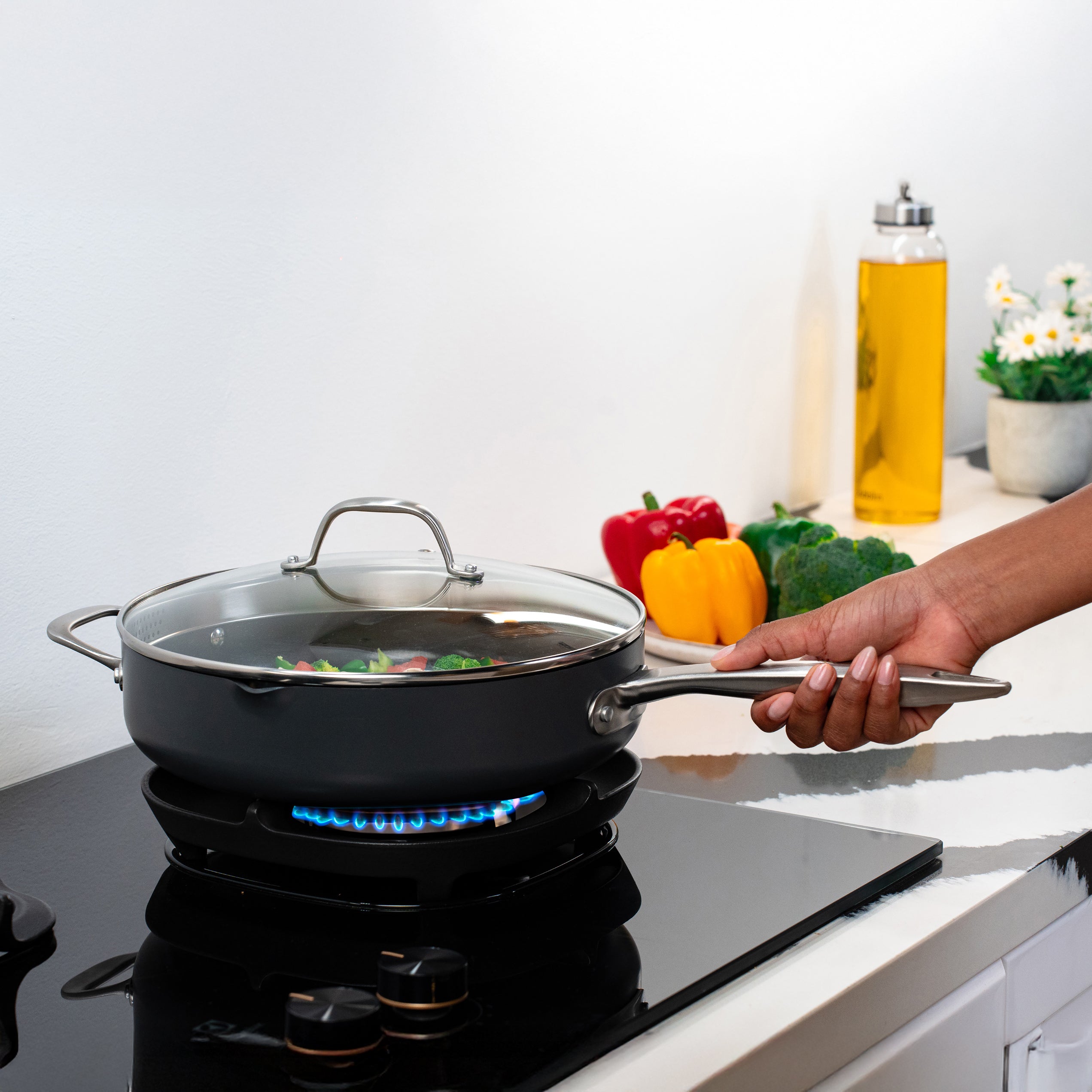 A hand holds the handle of a covered pan with vegetables cooking on a gas stove. In the background, bell peppers, broccoli, and a bottle of oil are on the counter near a small flowerpot.
