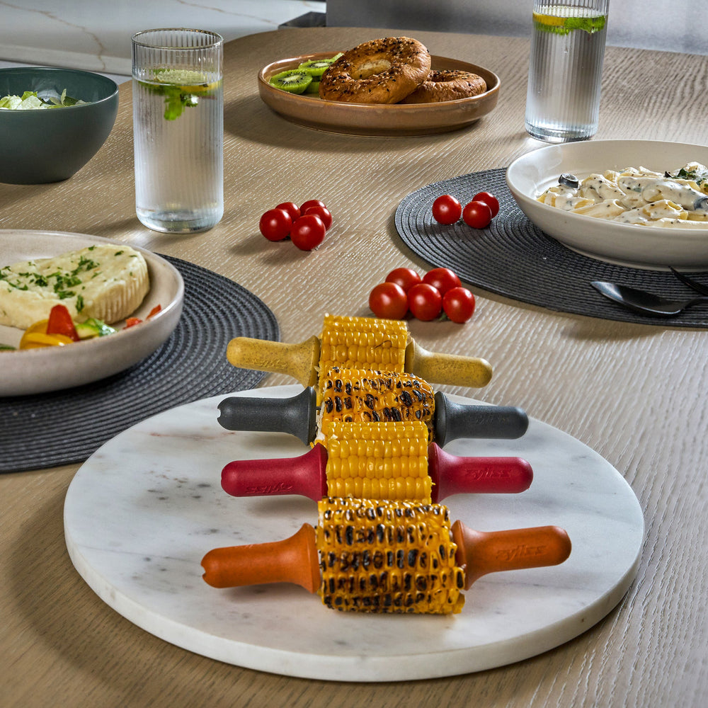The Zyliss Corn Holders 4 Piece Set features cylindrical holders with ergonomic handles in yellow, black, orange, and red, each with a notch at one end, shown vertically on a light background.
