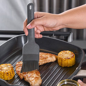A hand uses the Zyliss Silicone Pastry Brush to spread sauce on grilled chicken breast in a grill pan, with two grilled corn cobs beside the chicken on the stove.