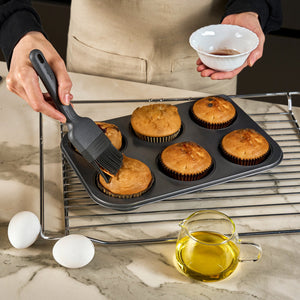 A person uses the Zyliss Silicone Pastry Brush to glaze muffins in a baking tray while holding a small bowl. The tray rests on a cooling rack, with two eggs and a pitcher of oil on the marble countertop nearby.