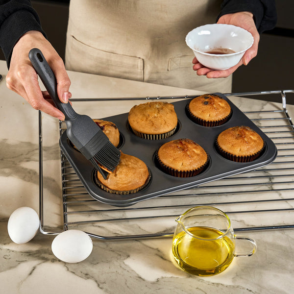 A person uses the Zyliss Silicone Pastry Brush to glaze muffins in a baking tray while holding a small bowl. The tray rests on a cooling rack, with two eggs and a pitcher of oil on the marble countertop nearby.
