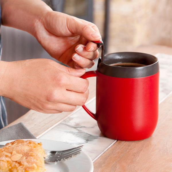A person holds the handle of a red mug filled with a hot beverage. A plate with a pastry and a fork sits on the wooden table in front of them.