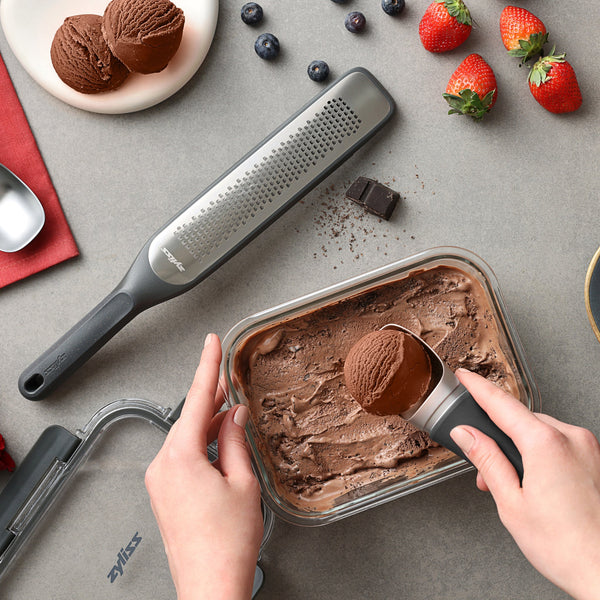 A person scoops chocolate ice cream from a container. Nearby are a grater, fresh berries, an ice cream scoop, and Zyliss Glass Containers for easy meal prep—all arranged on a gray surface with a red napkin. #1.05L/1.1qt