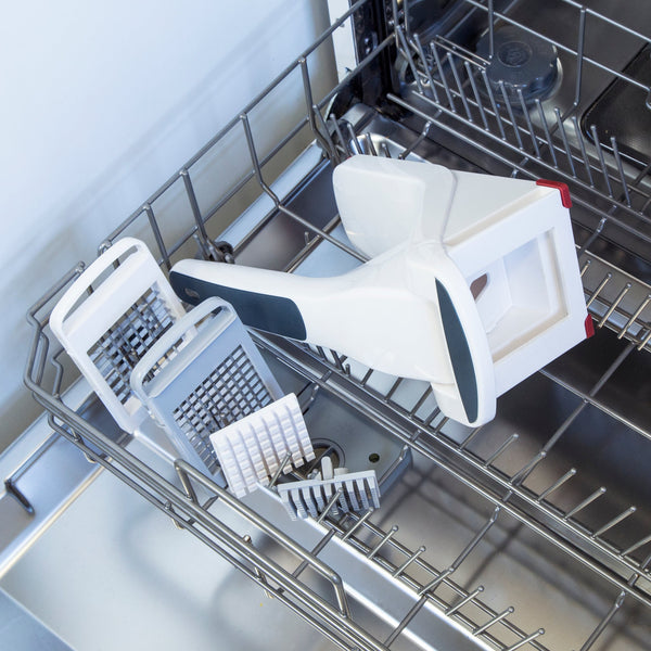 A disassembled white and black kitchen tool with several grater attachments is placed on the top rack of an open dishwasher.
