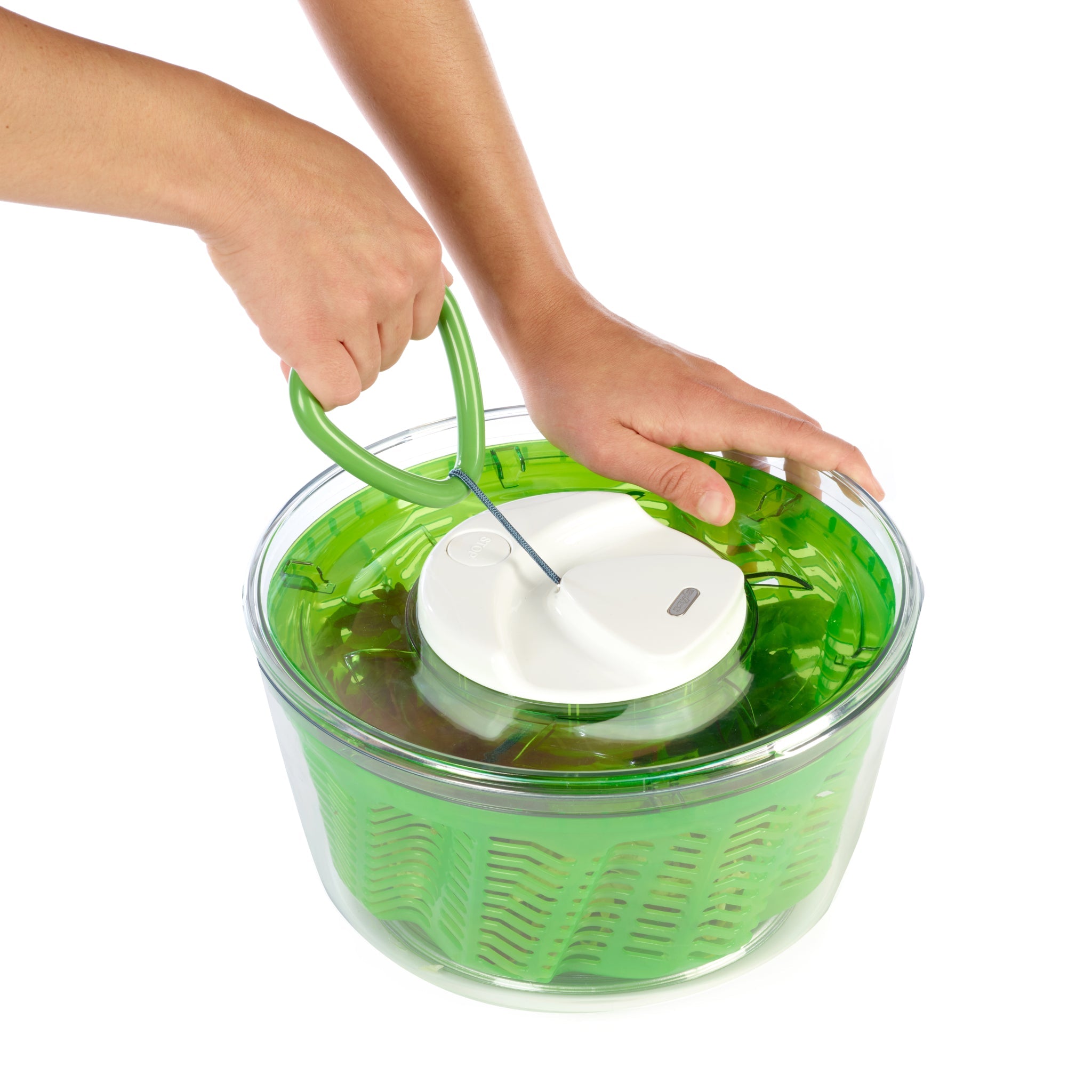 Hands using a green and clear salad spinner with a white lid to dry leafy greens inside a transparent bowl.