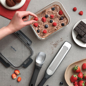 A hand places berries on chocolate ice cream in a glass container. Nearby are a chocolate bar, grater, ice cream scoop, lid, and strawberries on a gray surface with a red napkin and bowl.