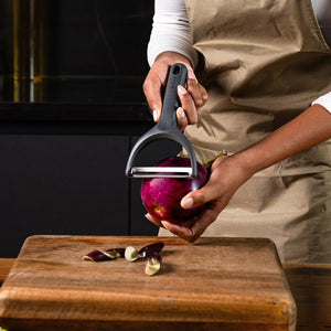 Wearing an apron, someone uses the Zyliss Wide Peeler with its extra-wide stainless steel blade to peel a purple eggplant over a wooden cutting board, leaving eggplant peels scattered on the board.