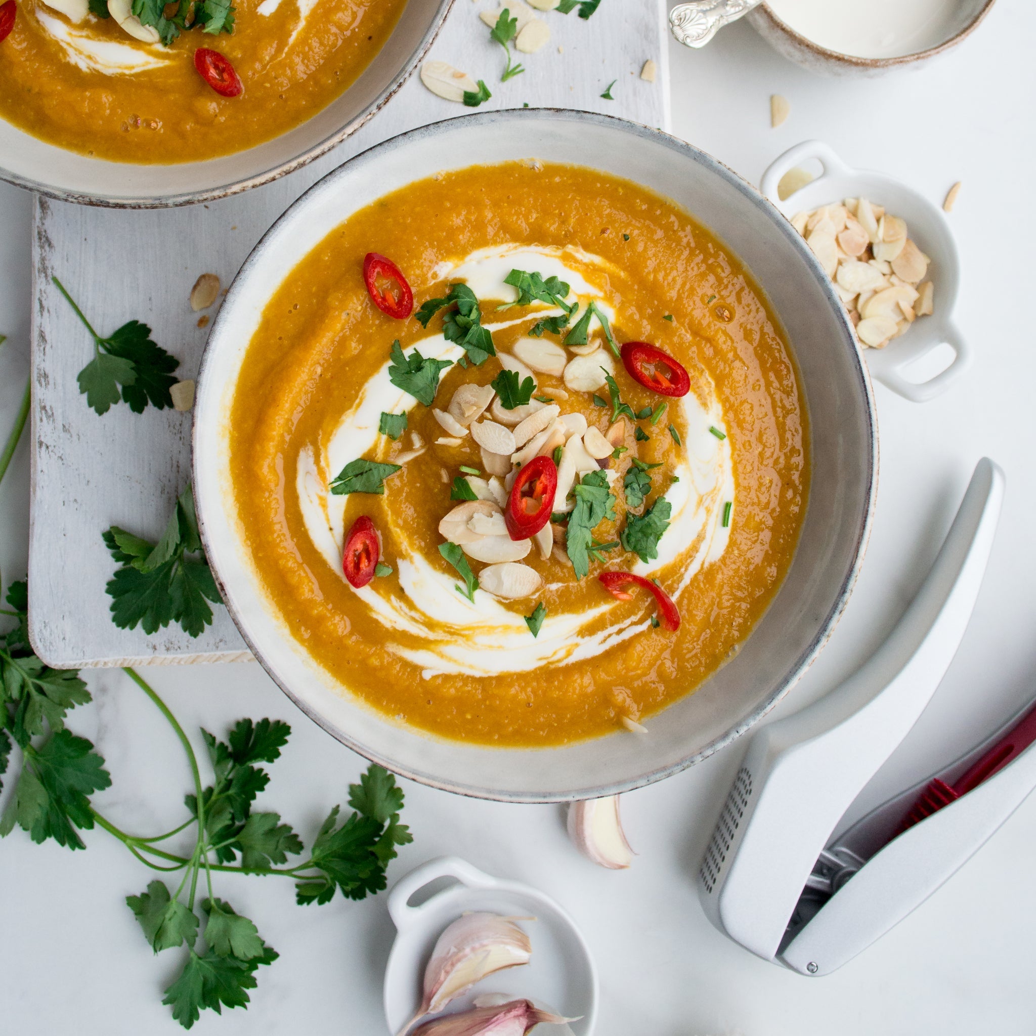 A bowl of orange soup with cream, chili, herbs, and almond flakes is surrounded by parsley, garlic, and the Zyliss Susi 3 Garlic Press featuring an ergonomic handle on a white surface. 
