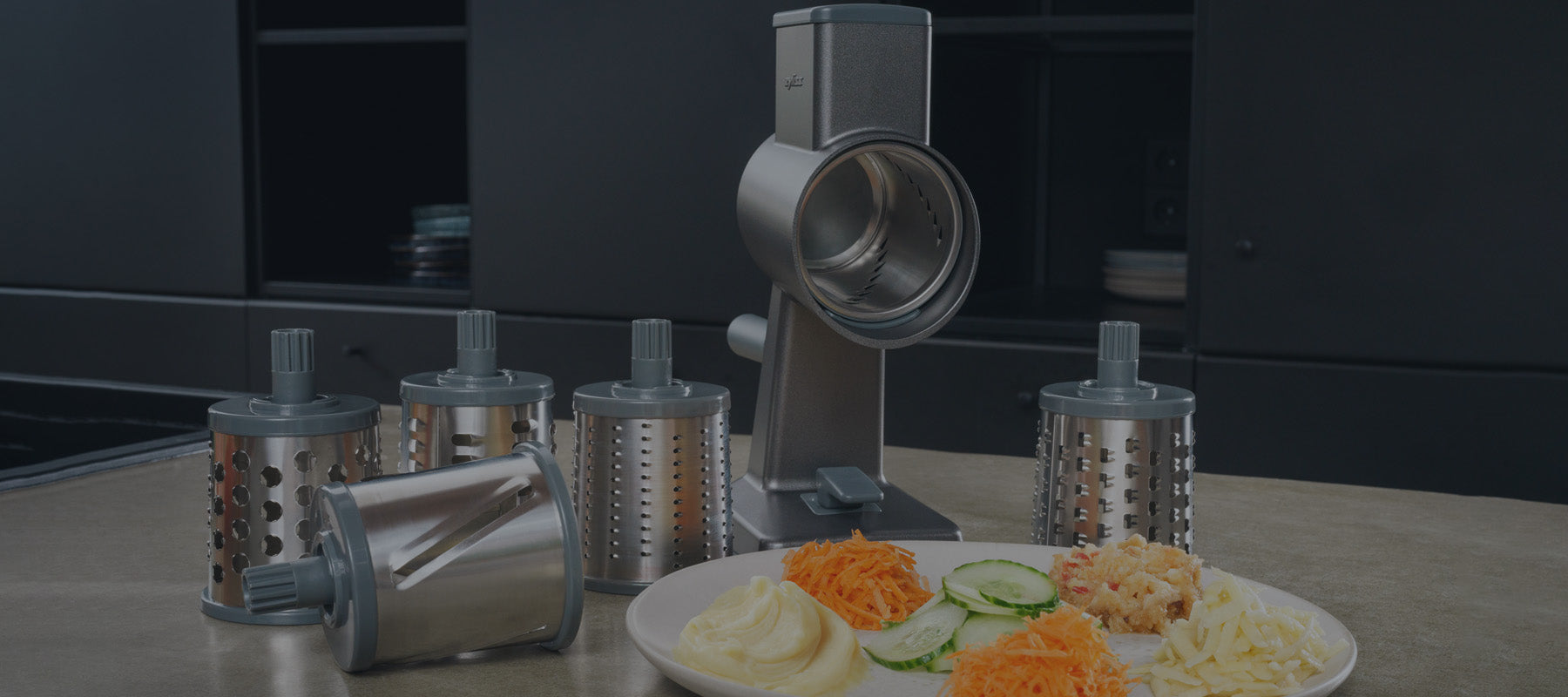 A stainless steel rotary grater with five interchangeable drum blades is displayed on a kitchen counter next to a plate with sliced cucumbers, grated carrots, shredded cheese, and mashed potatoes.