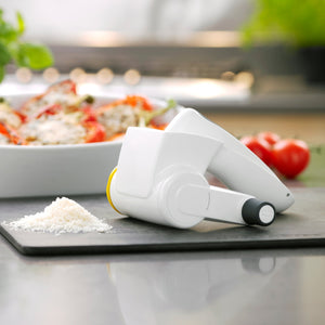 The Zyliss Classic Cheese Grater, featuring an ergonomic white handle, rests on a kitchen counter with grated cheese in front. A tasty dish and fresh tomatoes complete the inviting scene.