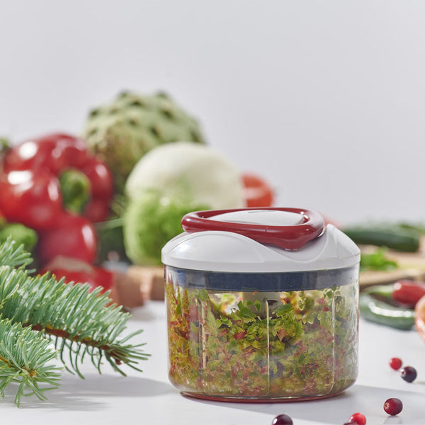 A manual food chopper filled with chopped vegetables sits on a table, surrounded by fresh produce such as bell peppers, an artichoke, and a sprig of pine, with a white background.