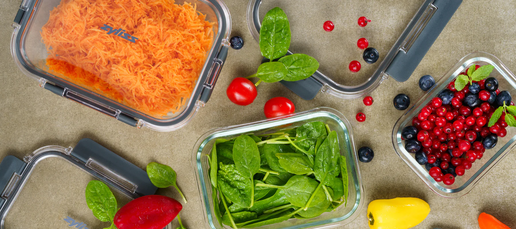 Glass containers filled with shredded carrots, fresh spinach, red currants, blueberries, and cherry tomatoes are arranged on a countertop, along with a yellow pepper and a piece of red beet.