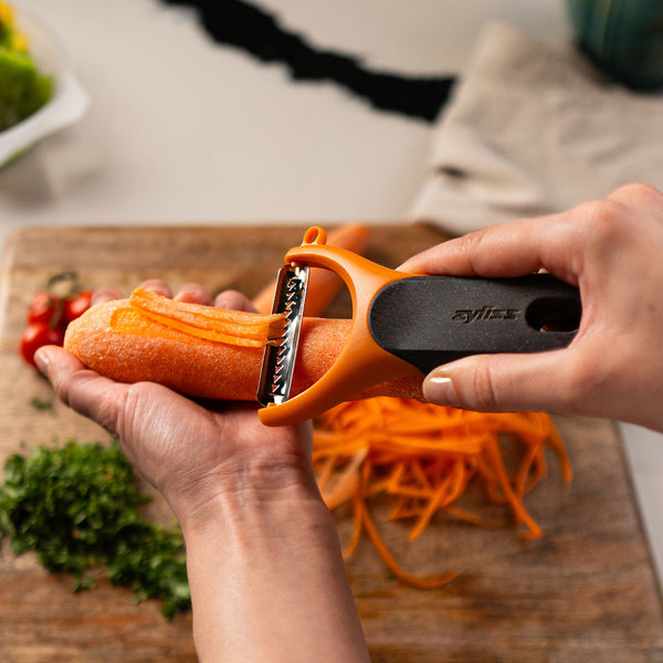 A person uses an orange vegetable peeler to julienne a carrot over a wooden cutting board, with sliced vegetables and chopped herbs visible in the background.