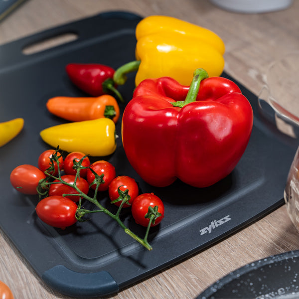 A black Zyliss cutting board with a red bell pepper, yellow bell pepper, small orange and yellow peppers, and a vine of cherry tomatoes on a wooden surface.